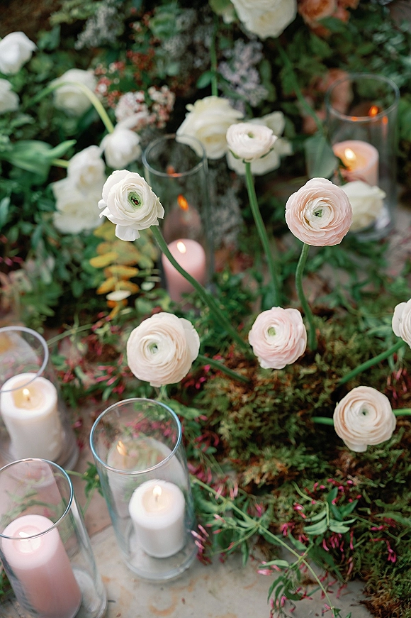 Wedding floral arrangement with ranunculus wedding flowers, greenery and moss around pillar candles in glass hurricane vases on a stone floor