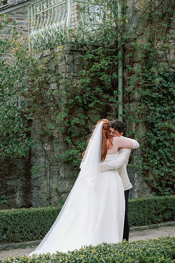 Couple portrait of bride and groom hug, her cathedral veil flowing behind a strapless gown by an ivy stone wall and balcony