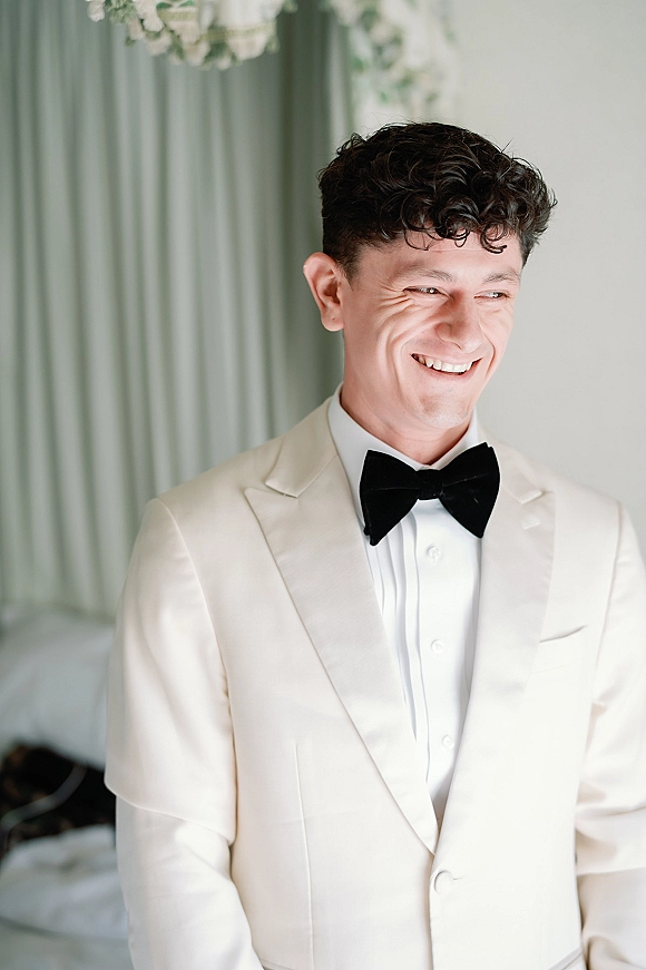 Groom portrait in a cream tuxedo jacket and black bow tie, looking away with a soft smile in a bright room with curtains and bed