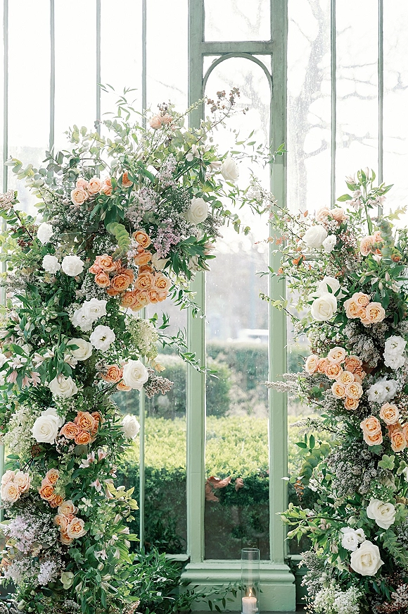 Wedding ceremony backdrop with an asymmetrical floral arch of peach and white roses, greenery, and a pillar candle before large windows