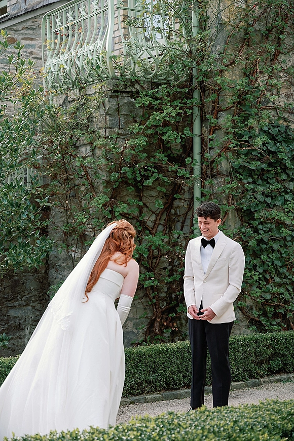 First look moment as bride in strapless gown and long veil approaches groom in white tuxedo by ivy stone wall and hedged path