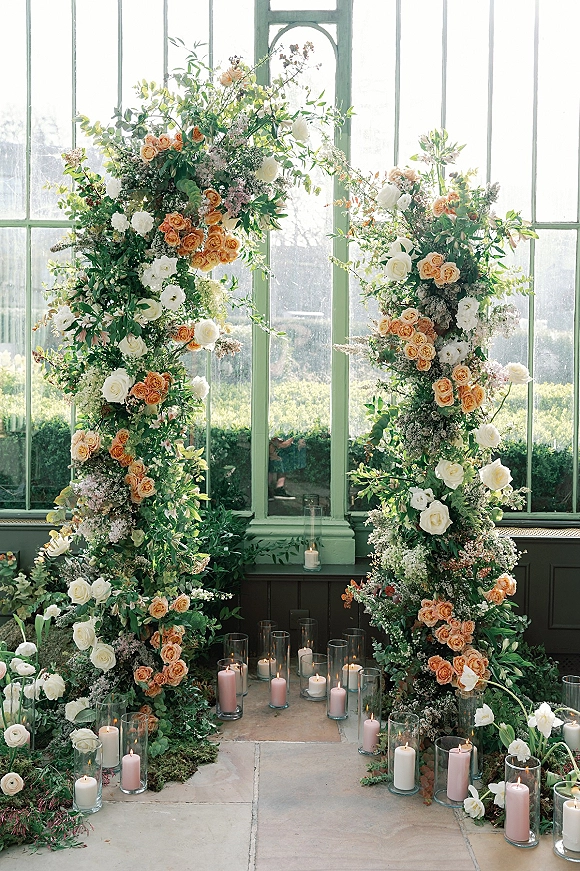 Wedding ceremony arch with floral wedding arch greenery, peach and white roses, and candle-lined glass hurricanes in a greenhouse setting