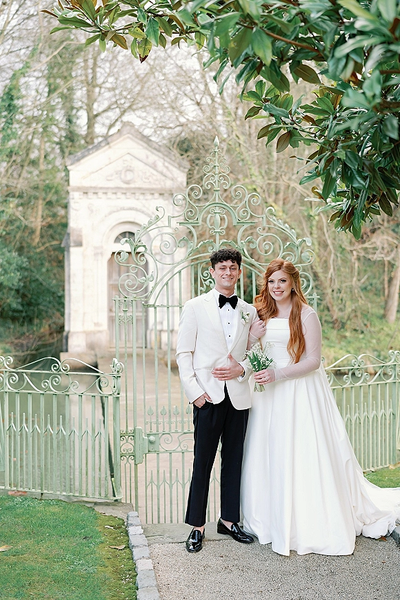 Couple portrait of bride in strapless dress and veil holding a white bouquet beside groom in tuxedo at a wrought iron gate in a garden estate