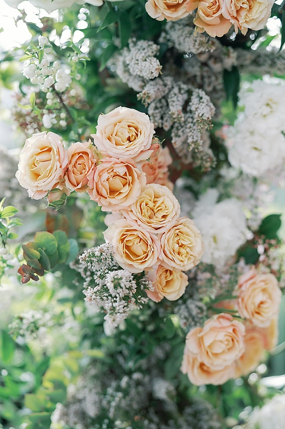 Wedding floral arch with peach roses and white blooms, baby’s breath and lush greenery, set against soft garden foliage outdoors