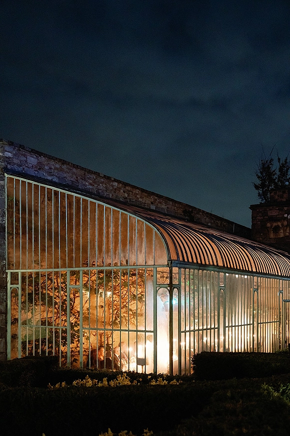 Greenhouse wedding reception with a long table set under string lights and candlelit glow inside a glass-and-metal conservatory at night