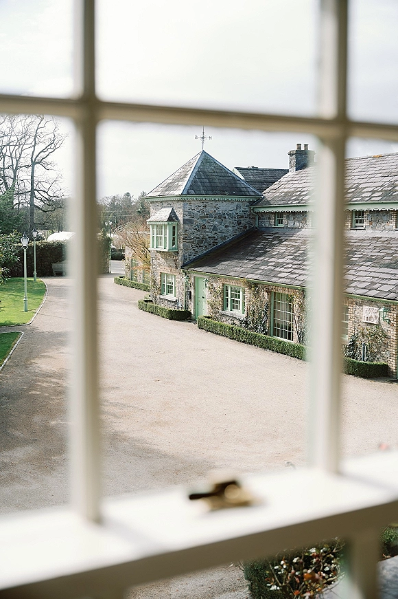 Wedding venue exterior of a stone manor wedding venue with mint green windows, slate roof, ivy vines, and a hedge-lined driveway