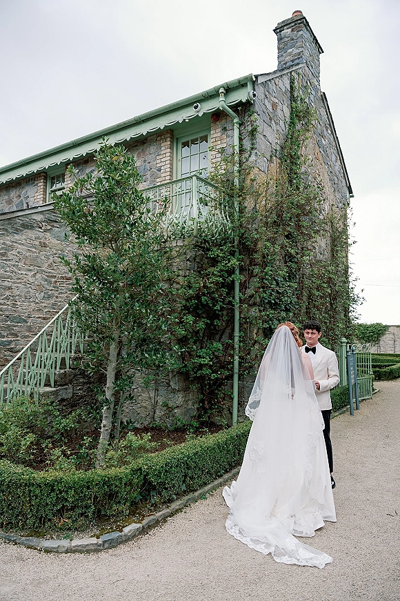 Wedding couple portrait of bride and groom facing each other, her long lace veil and dress train on a gravel path by an ivy stone wall