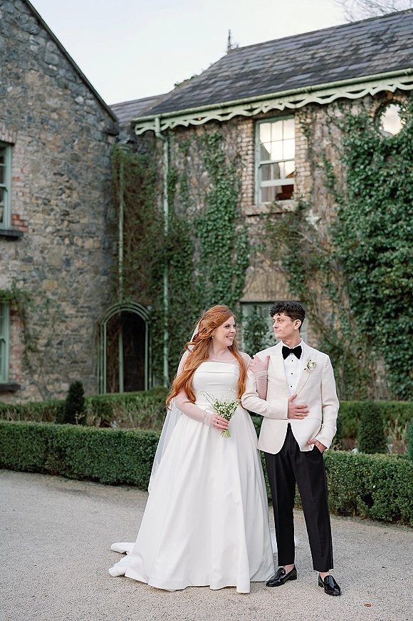Couple portrait of bride in strapless wedding dress and veil holding a small bouquet, beside groom in white tux by ivy stone courtyard