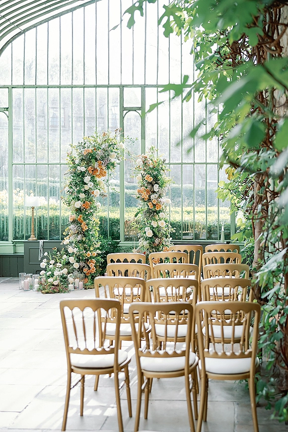 Ceremony setup in a glass greenhouse wedding ceremony with floral pillars of roses and greenery, wooden chairs, and pillar candles on stone floor aisle