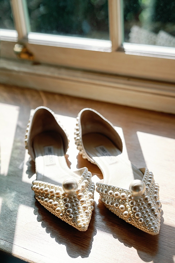 Bridal shoes with pearl embellishments, pointed-toe flats resting on a wooden surface by a sunlit window with soft greenery bokeh