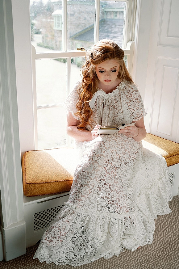 Bridal portrait of a bride getting ready in a lace gown, seated by a window, holding a notebook and showing her ring in soft light
