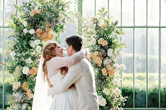 Wedding kiss portrait of bride and groom kissing beneath a floral arch with roses and greenery, veil draped in a greenhouse garden setting