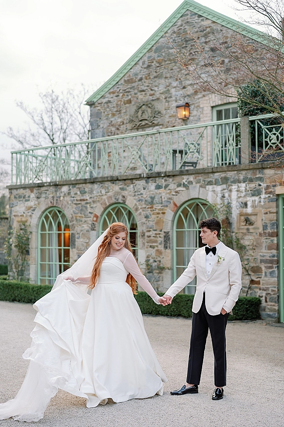Couple portrait of bride and groom holding hands, her veil and gown flowing as they walk in a courtyard by an arched stone facade