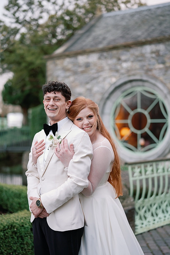 Couple portrait with bride hugging groom from behind, his white tuxedo jacket and her long veil set against a stone building with round window