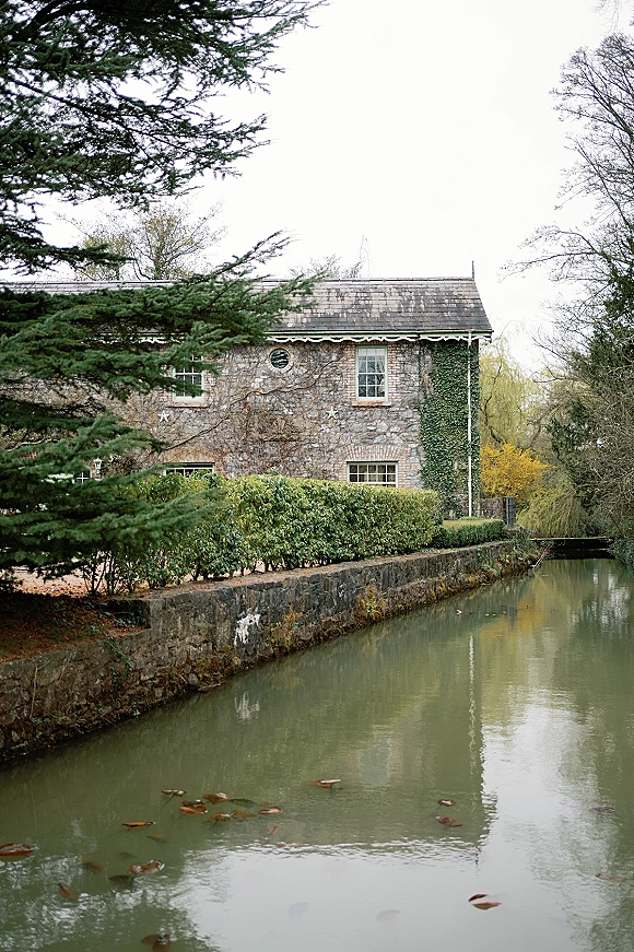 Wedding venue exterior of an ivy-covered stone manor with slate roof and windows, framed by hedges beside a canal under overcast sky