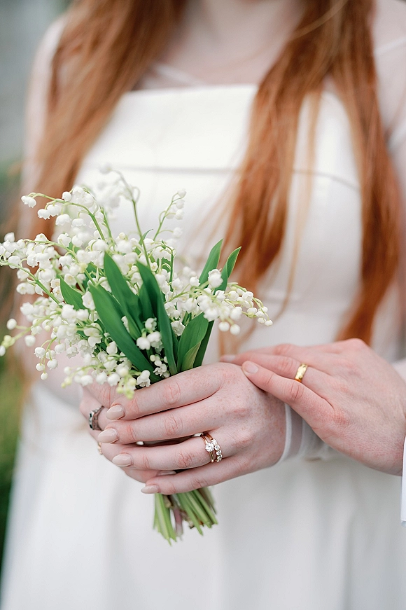 Bridal bouquet close-up of a lily of the valley bouquet in manicured hands, engagement ring and wedding band visible against blurred greenery