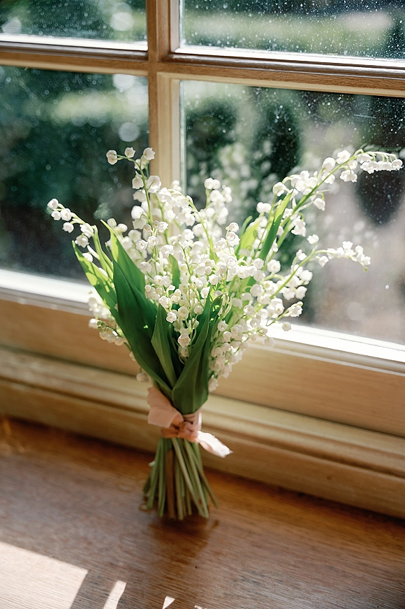 Lily of the valley bouquet with ribbon wrap and green leaves resting on a sunlit wooden windowsill with blurred greenery beyond