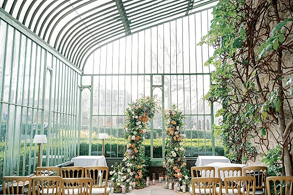 Ceremony setup in a glass greenhouse wedding ceremony with floral pillars, roses and greenery, and candlelit aisle beside white chairs