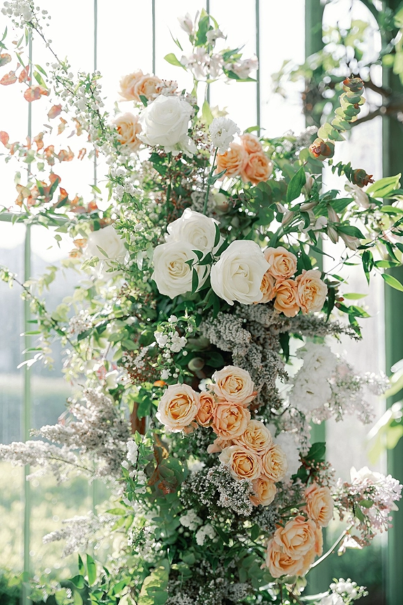 Wedding floral arrangement of peach and white roses with greenery and eucalyptus accents, glowing in sunlight by greenhouse glass windows