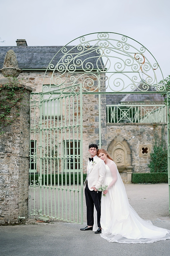 Couple portrait of bride hugging groom in a white tuxedo and holding a bouquet, framed by a wrought iron gate in a stone courtyard