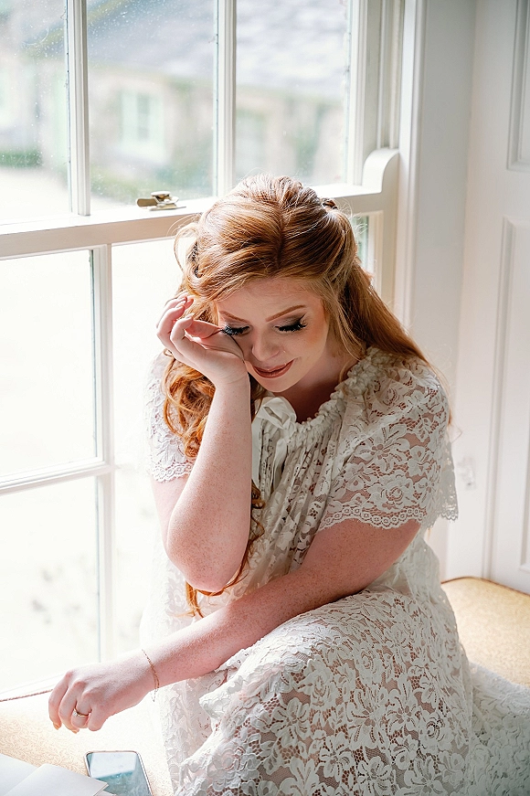 Bridal portrait of a bride sitting by window in a lace wedding dress, looking down with bracelet and wedding ring in soft light indoors