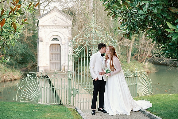 Wedding kiss portrait of bride and groom kissing, her veil and bouquet against a wrought iron gate in an estate garden setting