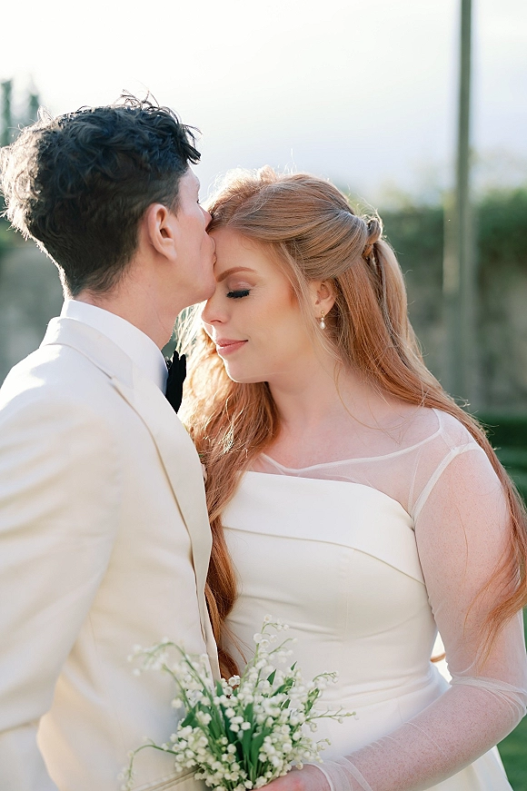 Wedding couple portrait with a groom kissing the bride’s forehead as she holds a lily of the valley bouquet in soft outdoor greenery light