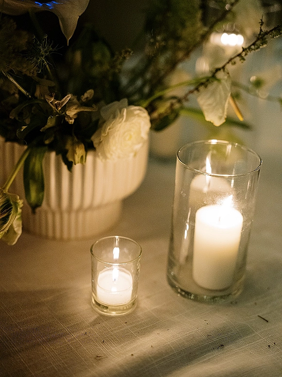 Wedding table candles glow in glass hurricane holders with white florals and greenery on a linen tablecloth, reception lighting softly blurred