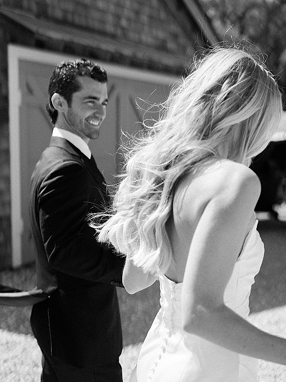 First look moment as bride in strapless gown and veil approaches groom in tuxedo from behind in a sunlit outdoor courtyard