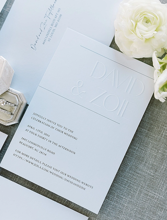 Wedding invitation suite with embossed wedding invitation, RSVP card, envelope, rings, and white flowers on gray linen fabric flat lay