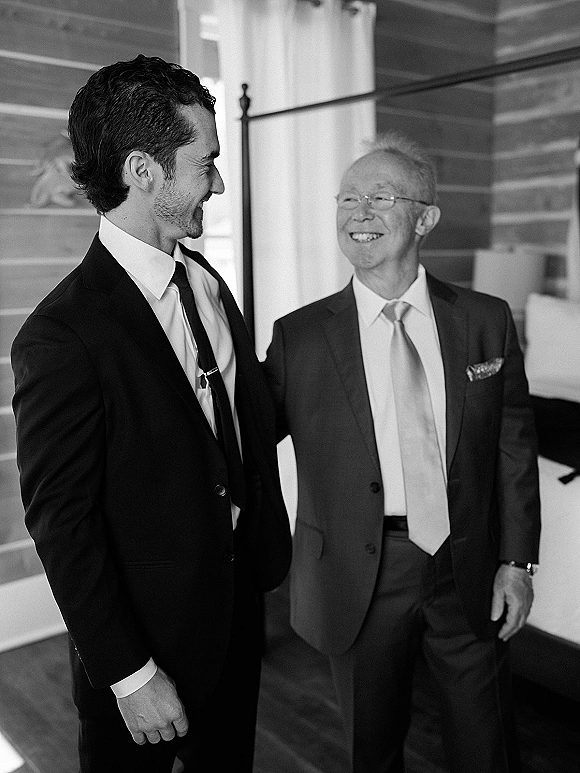 Groom with father in suits, sharing a father son wedding photo moment indoors, adjusting tie clip by wood-paneled wall and bed