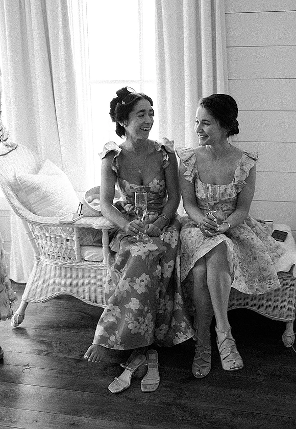 Bridesmaids portrait of friends laughing in floral bridesmaid dresses, holding champagne flutes on a wicker bench by a window with sheer curtains