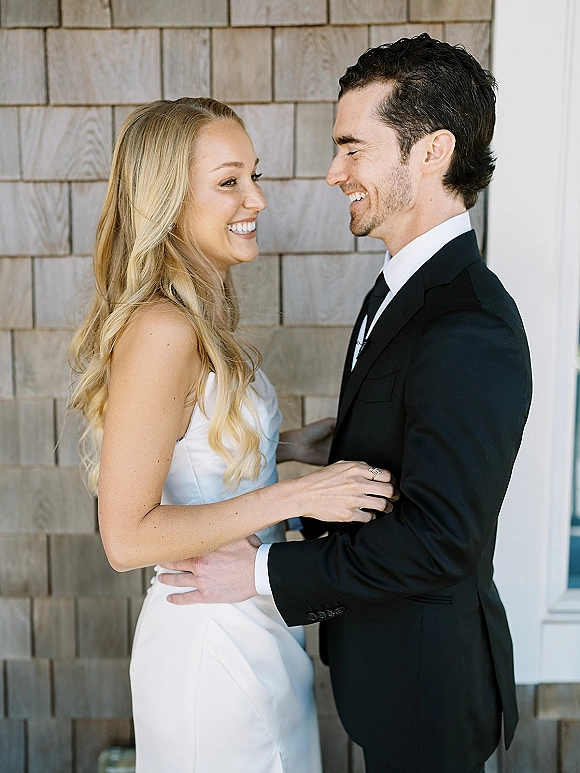 Couple portrait of bride and groom smiling face to face, holding each other as she touches his lapel by a shingle wall doorway trim