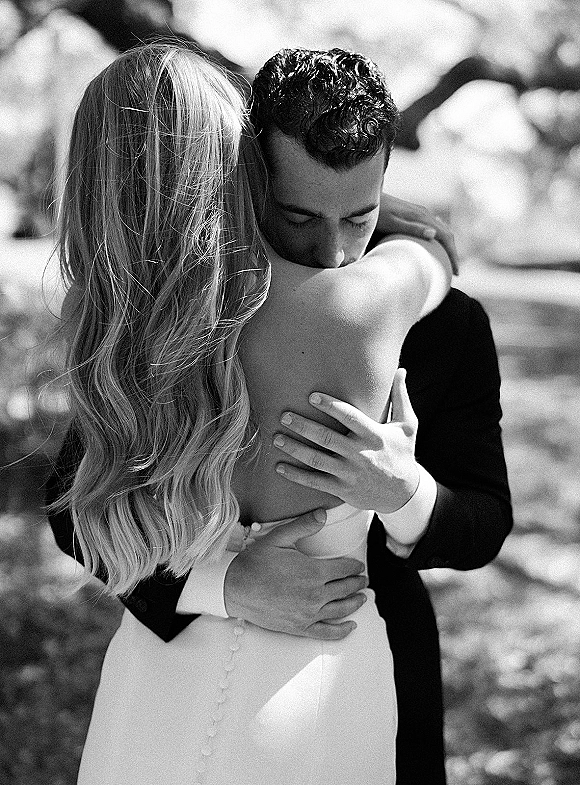 Wedding couple portrait of bride and groom hugging, showing a strapless button-back gown and long wavy hair under dappled tree shade