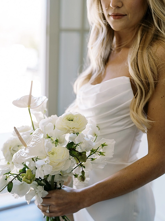 Bridal portrait of a bride holding bouquet of white ranunculus, anthurium and sweet peas, in window light, showing her engagement ring