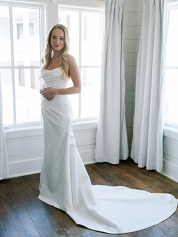 Bridal portrait of a bride by window in a strapless wedding dress with a long train, wavy hair, and engagement ring in soft natural light