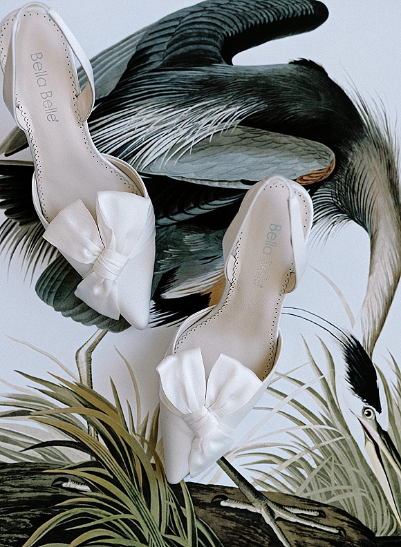 Bridal shoes in white satin heels with bow details and scalloped trim, styled on a botanical bird illustration flatlay