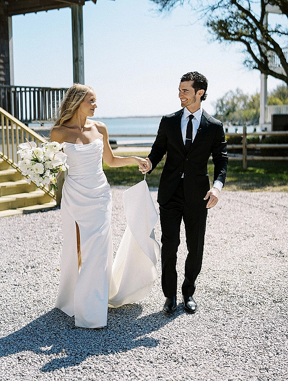 Couple portrait of bride and groom walking hand in hand, she holds a white bouquet on a waterfront gravel path, dress train flowing