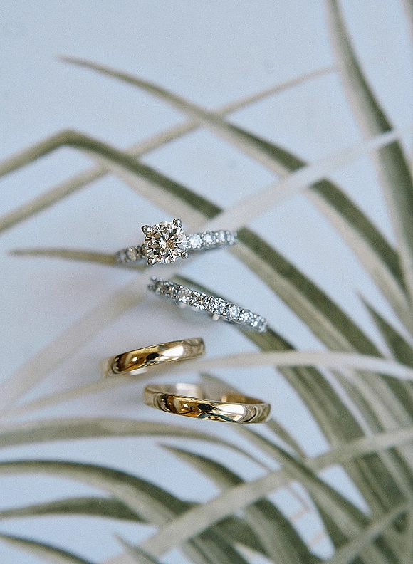Wedding rings flat lay with engagement ring close up, diamond solitaire and gold bands beside a palm leaf on a white surface