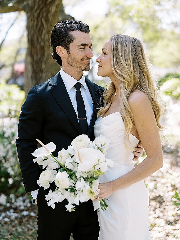 Couple portrait of bride and groom embracing, bride in strapless dress holding white calla lily bouquet in a sunlit garden setting