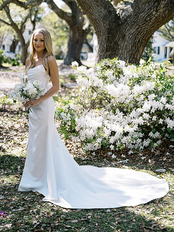 Bridal portrait of a bride holding bouquet of white calla lilies in a strapless gown with a long train in a garden by flowering shrubs