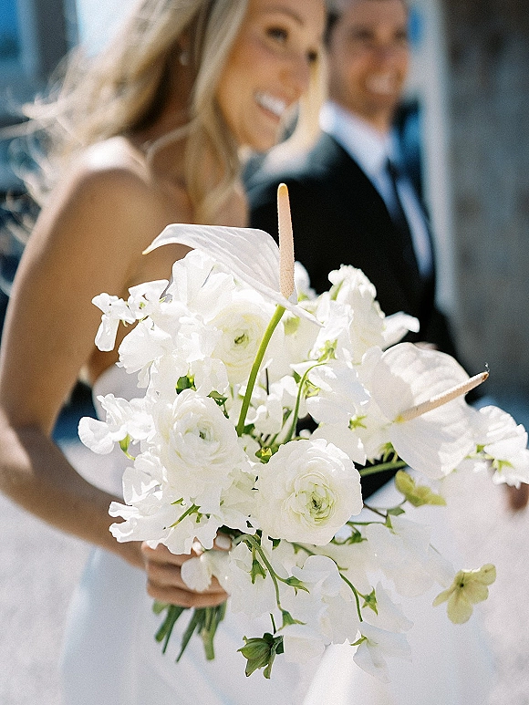 Bridal bouquet of white wedding bouquet blooms with anthurium, ranunculus, and sweet pea, held by bride and groom in sunlight by columns