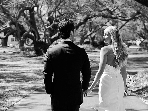 Couple portrait in a black and white wedding photo of bride and groom walking away holding hands on a tree-lined walkway, bouquet in hand