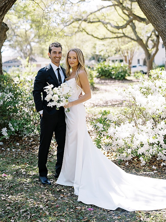 Couple portrait of bride in a strapless gown with long train and groom in black tuxedo holding a white bouquet in a sunlit garden