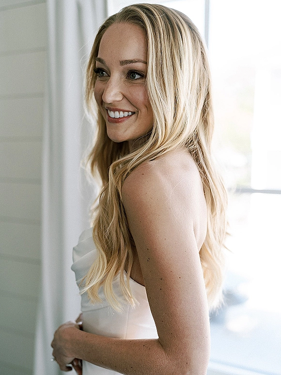 Bridal portrait of a smiling bride in side profile, wearing a strapless wedding dress, lit by soft window light against a white wall
