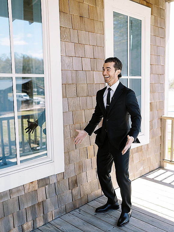 Groom portrait in a black suit with white dress shirt, black tie, and tie bar, smiling on a porch by windows and railing
