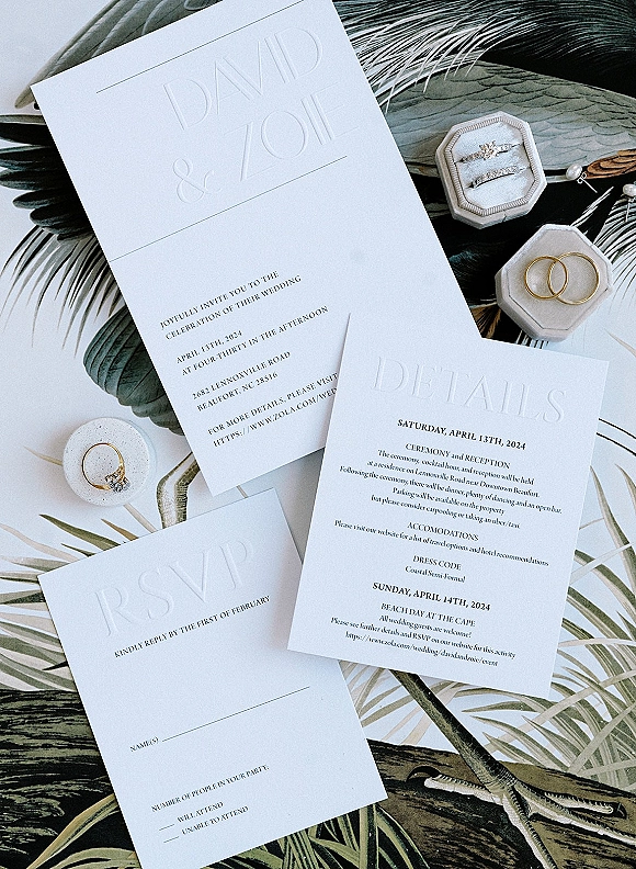 Invitation suite with embossed wedding invitations, RSVP and details cards, plus rings in a velvet box on a tropical leaf backdrop