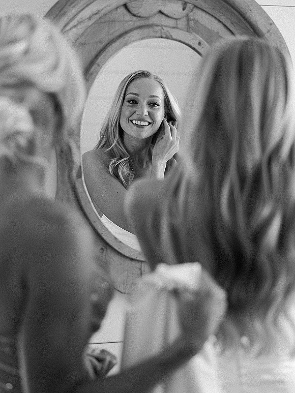 Bridal getting ready as bride in mirror adjusts earrings, hair down in white lingerie by a vanity, round mirror reflecting her smile