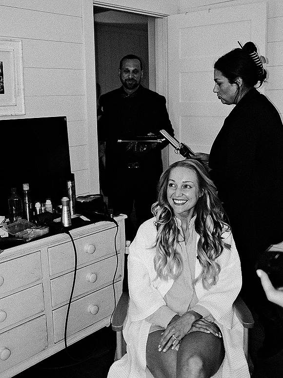 Bridal getting ready as bride hair styling with hair clips and curling iron, seated in a robe by a mirror in a bedroom
