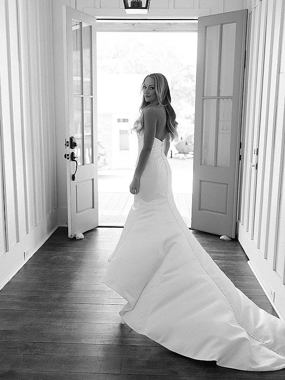 Bridal portrait of a bride looking back in a strapless wedding dress with long train and button back, framed by open double doors and soft light